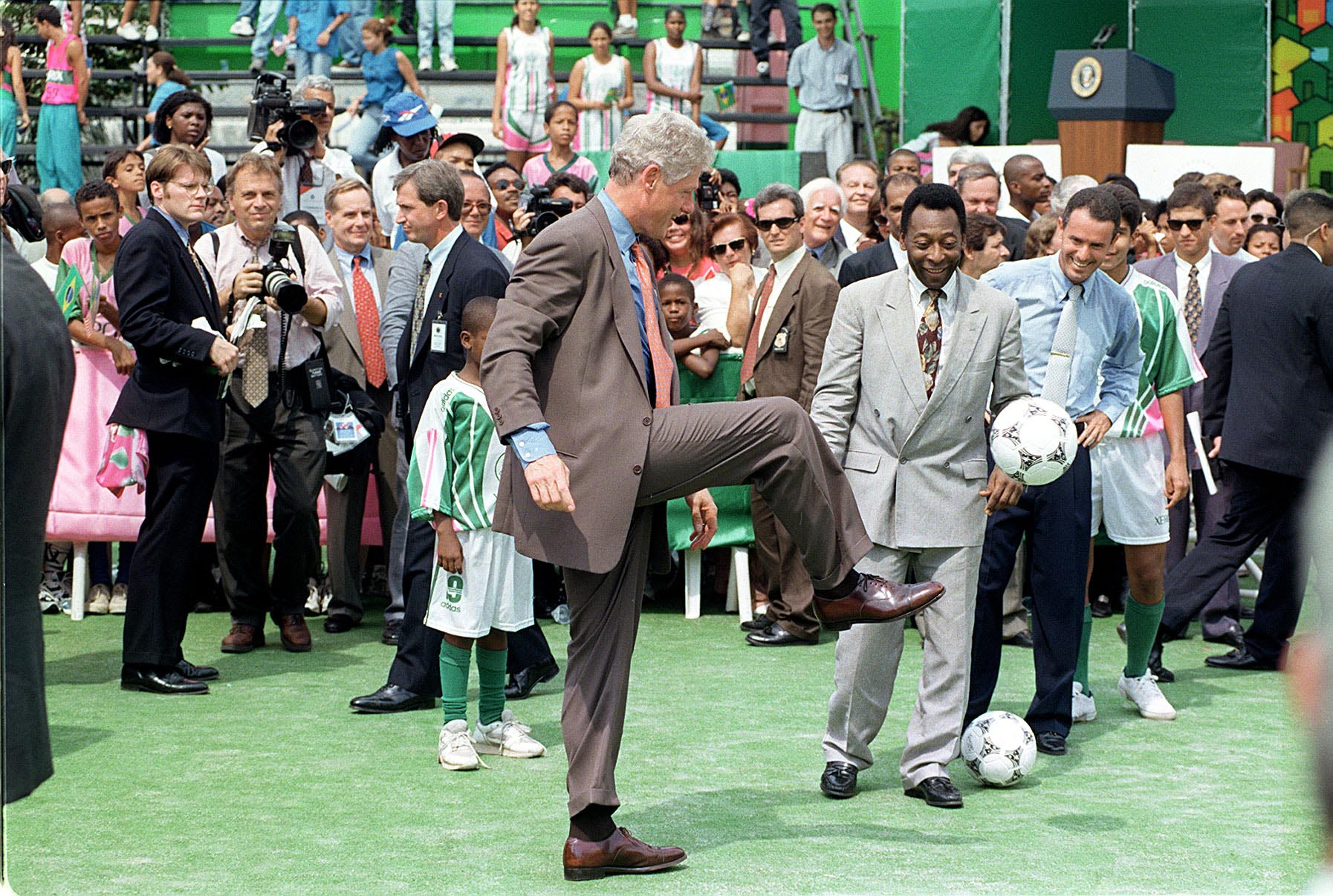 Pelé (de terno claro) acompanha o presidente norte-americano Bill Clinton (de terno marrom), durante sua visita à quadra da escola de samba Estação Primeira de Mangueira, no Rio de Janeiro, 1997 - Foto: Raimundo Valentim/Estadão Conteúdo.